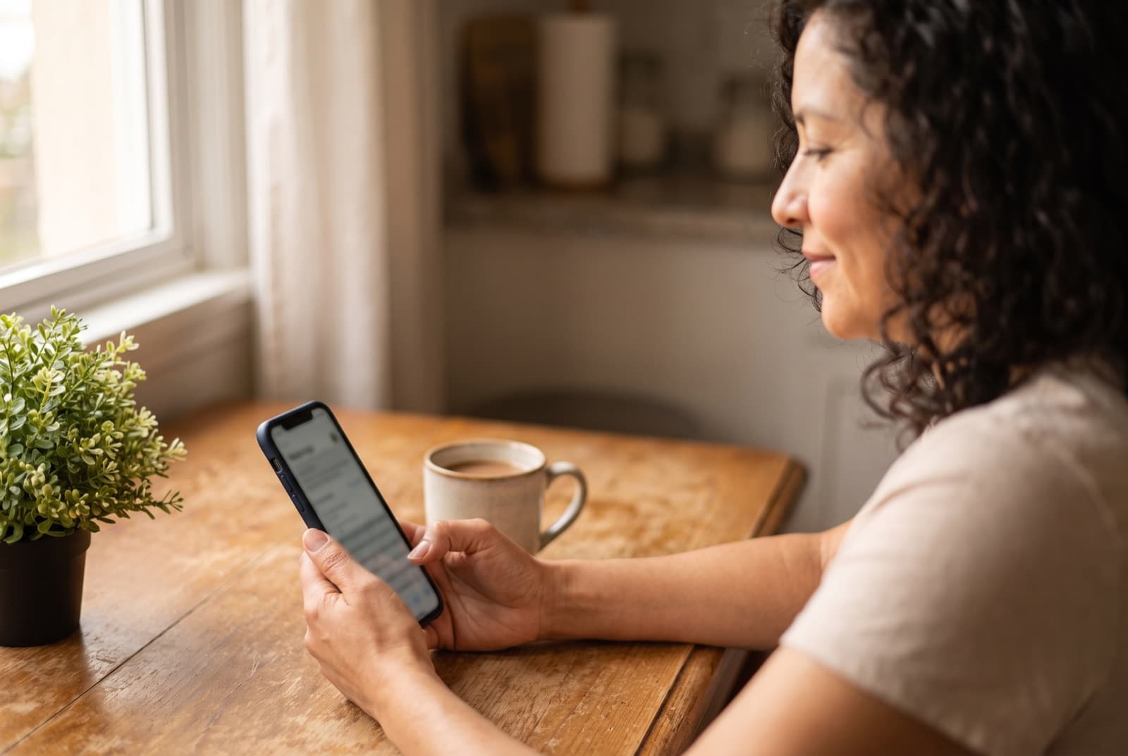 Mujer en la mesa de su cocina mirando el móvil con una taza de café al lado